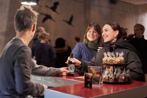 Laechelnde Frau bestellt etwas an der Bar bei der Werkschau des Studiengangs Film und Motion Design – Eventfotograf Hamburg