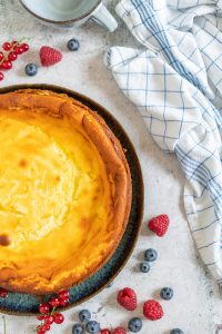 Kaesekuchen mit Himbeeren und Blaubeeren und Johannisbeeren neben einem Tuch und einer Tasse – Foodfotograf Hamburg