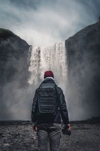 Ein Mann mit Rucksack und Kamera steht vor einem Wasserfall auf Island – Landschaftsfotograf Hamburg