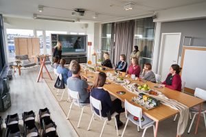 Viele Personen sitzen an einem langen Tisch in einem Seminarraum und hoeren einen Vortrag